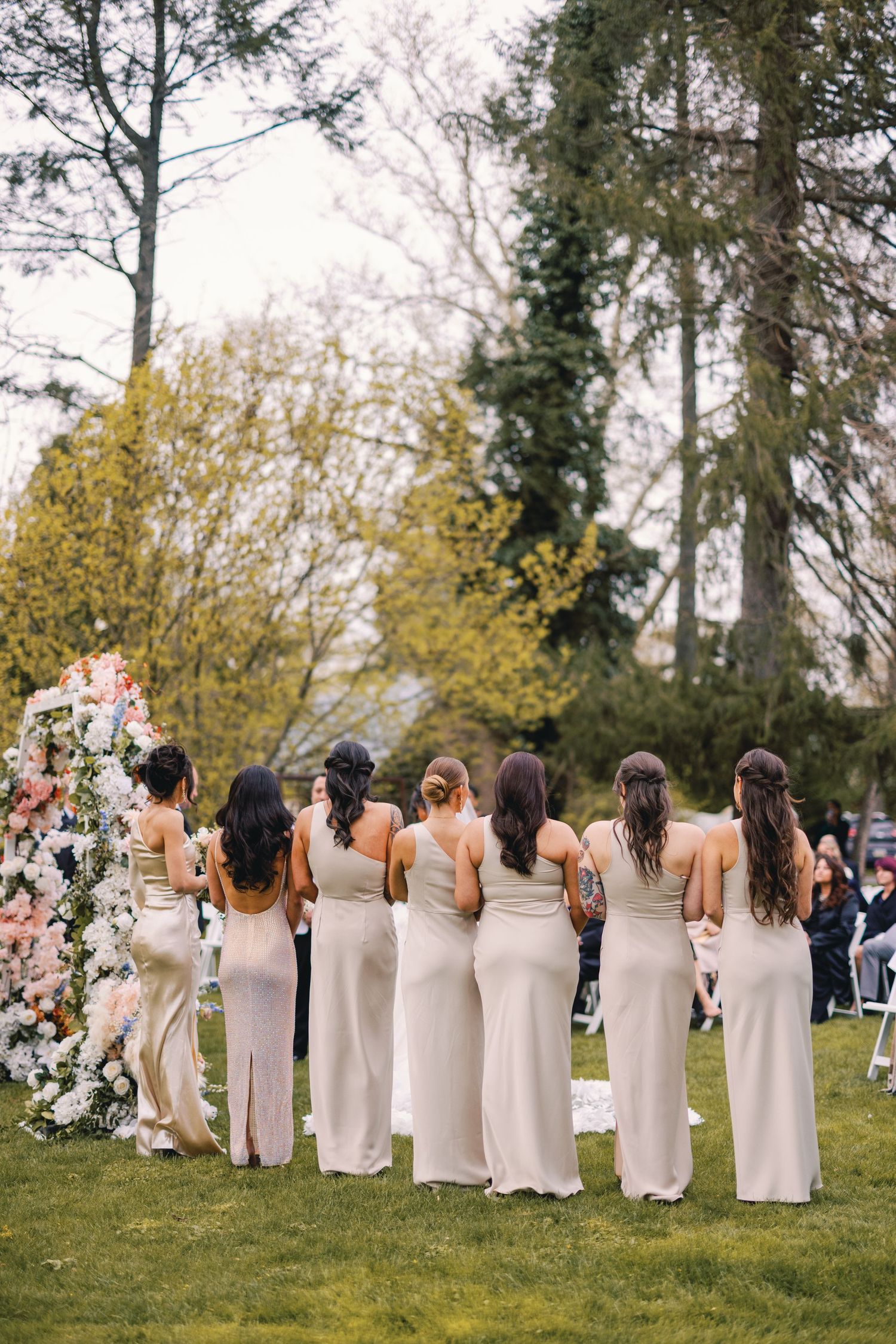 Six bridesmaids in champagne gowns stand before floral arch at outdoor garden ceremony.