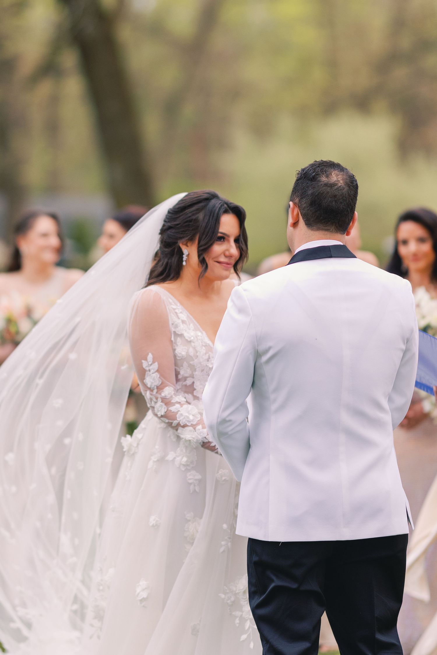 Bride smiles at groom during outdoor wedding ceremony with flowing veil and lace gown.