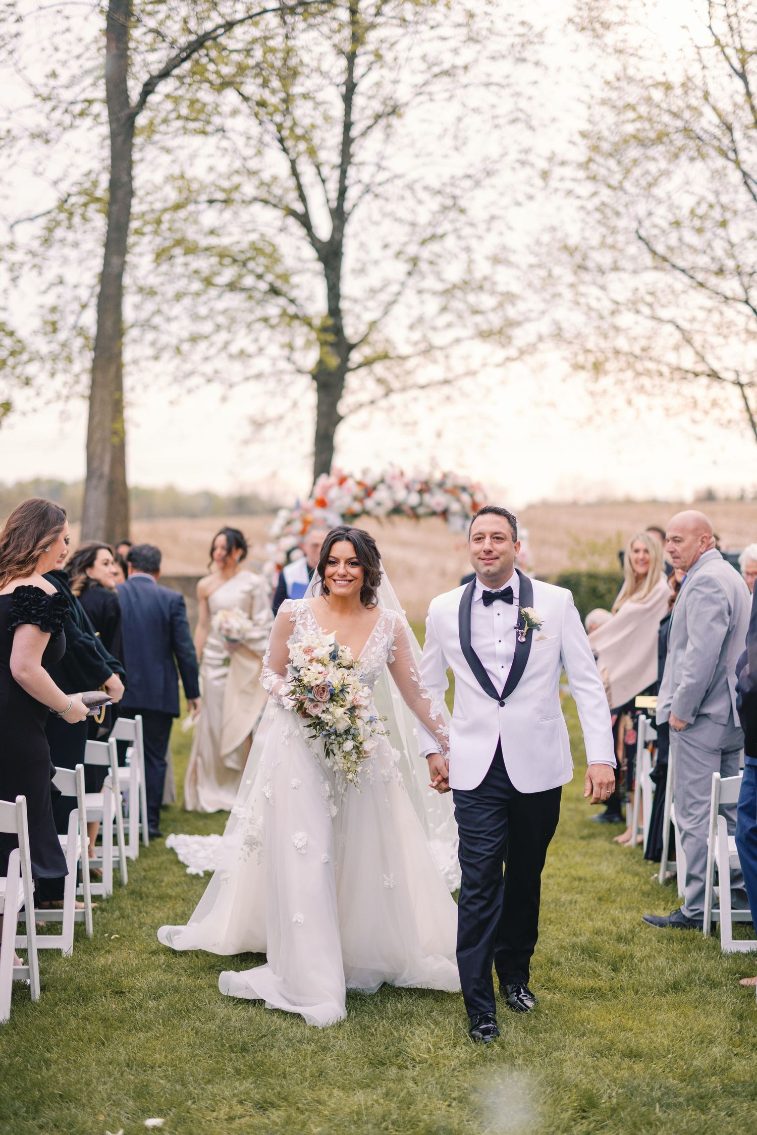 Smiling bride and groom walk hand in hand down outdoor wedding ceremony aisle.