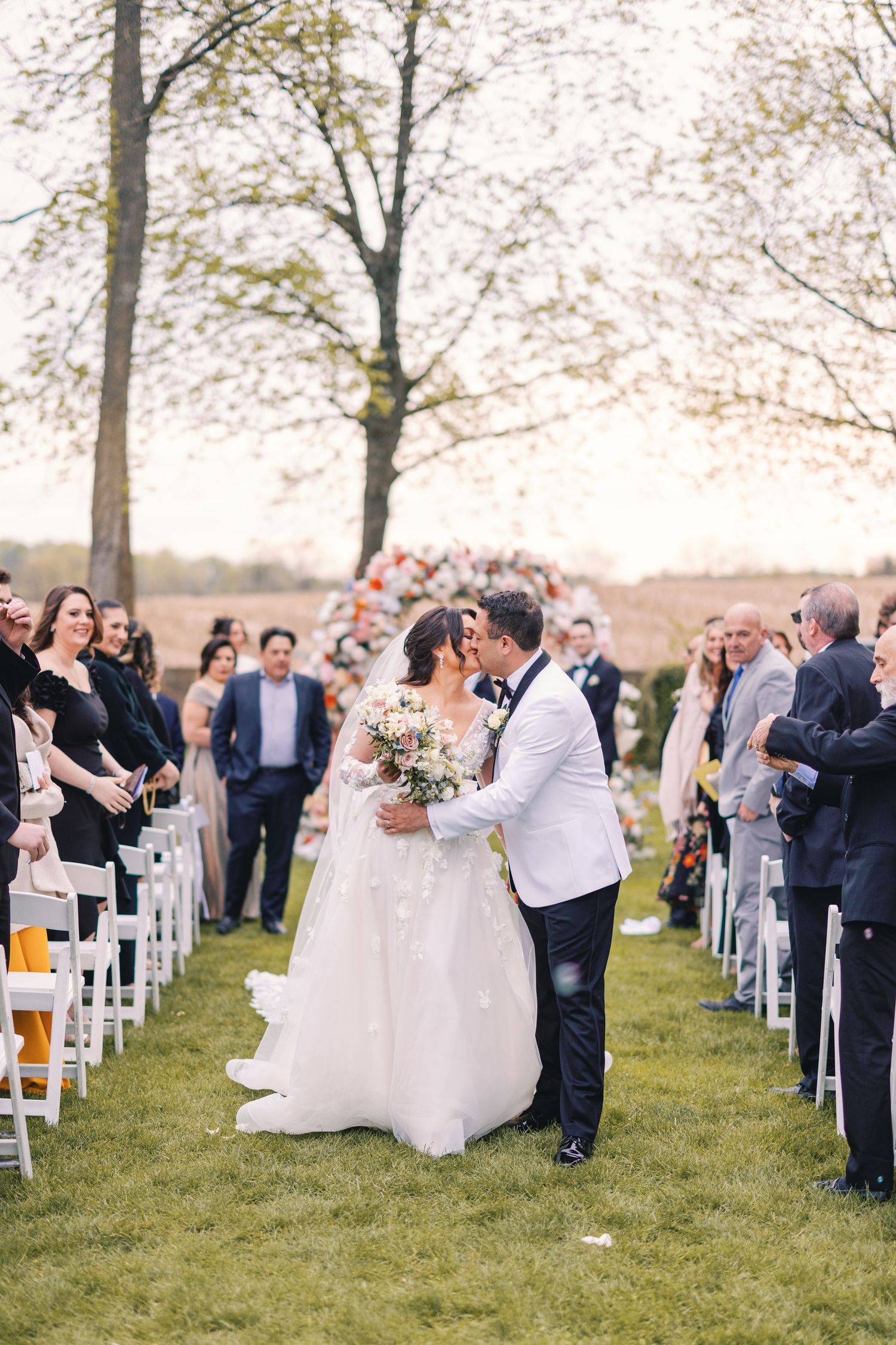 Bride and groom share first kiss at outdoor wedding ceremony with floral arch and white chairs.