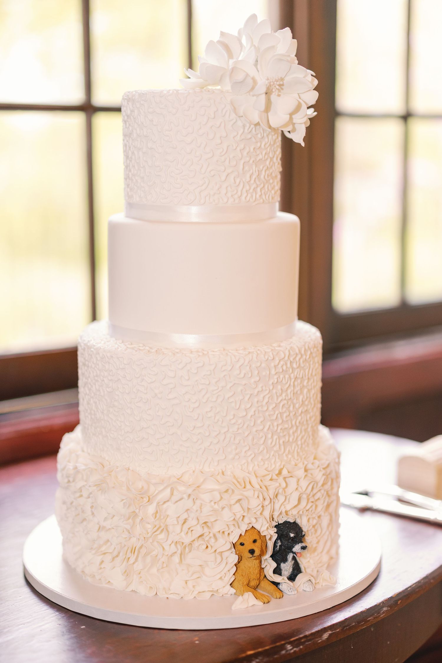 Four-tier white wedding cake with textured ruffles, floral topper, and cute dog cake toppers.