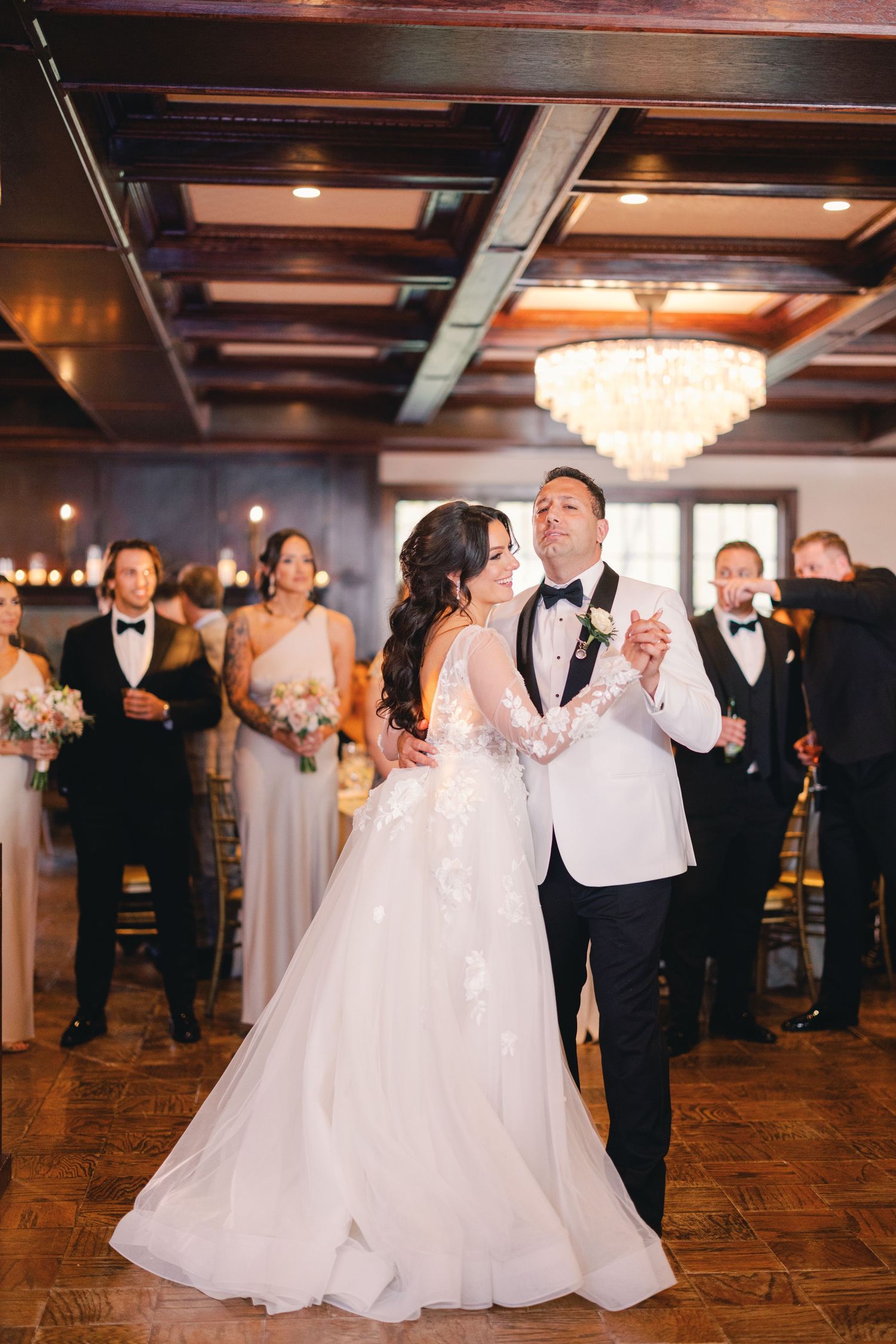 Bride and groom share romantic first dance under chandelier in elegant wood-paneled reception hall.