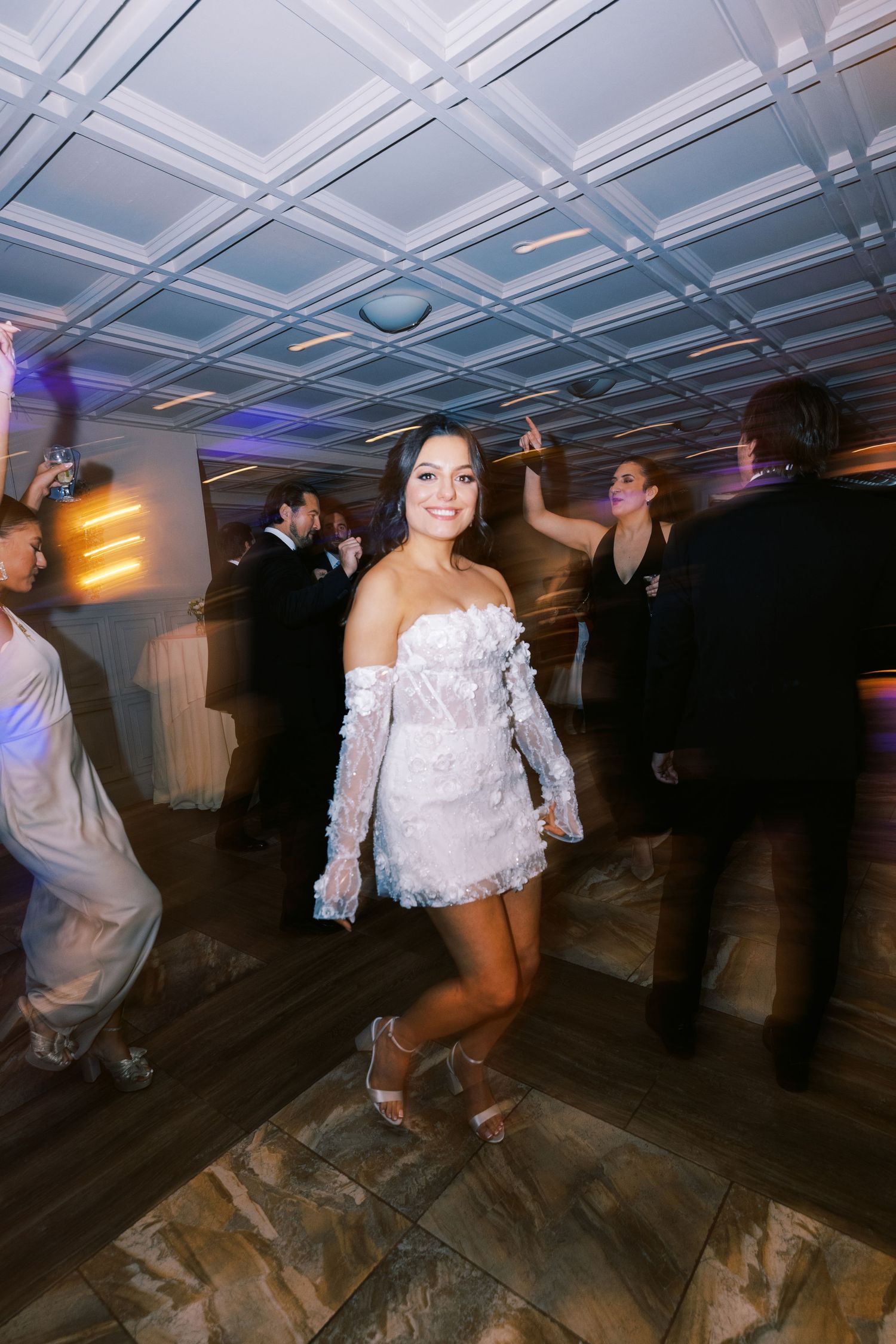 Bride in short white floral mini dress smiles while dancing at wedding reception party.