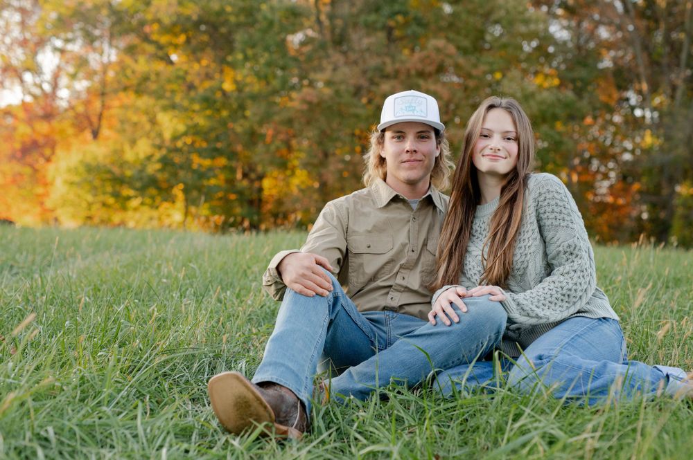A couple in casual attire sits together on grass during an autumn outdoor photoshoot.