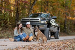 A couple sits with their dogs in front of a lifted black Dodge Ram truck surrounded by fall foliage.