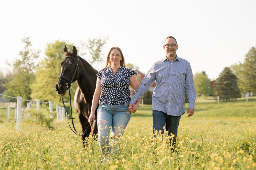 A couple walks alongside a black horse in a sunny pasture with white fencing.