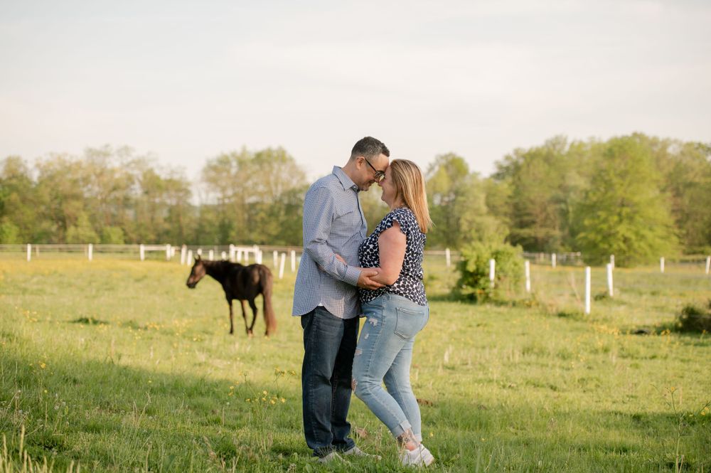 A romantic couple embraces in a pastoral field with horses grazing in the background.