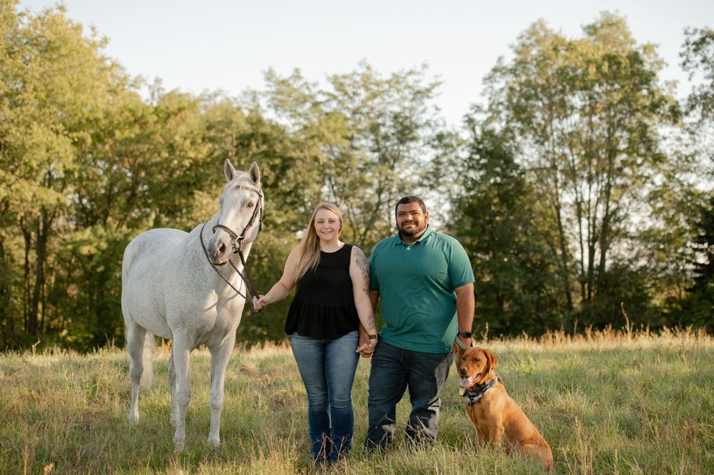 A couple poses with their white horse and brown dog in a sunny field with trees in the background.