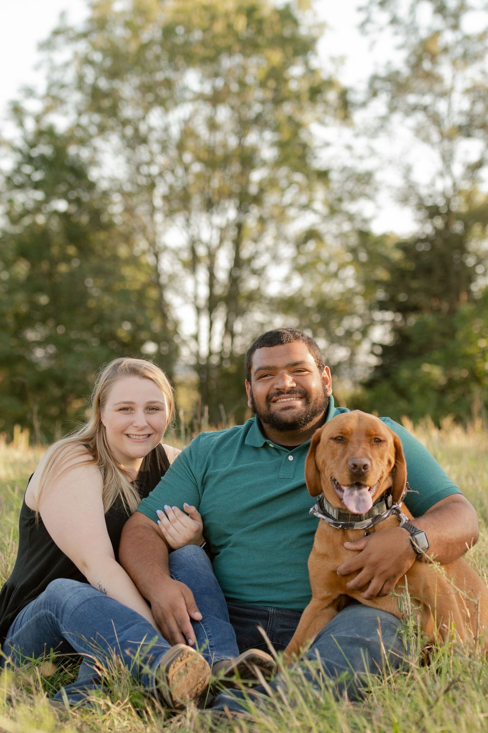A couple sits in a grassy field with their happy brown dog during golden hour.