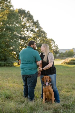 A couple embraces while their brown dog sits beside them in a sunny meadow.