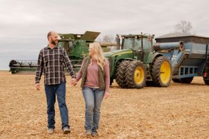 Couple walking hand-in-hand through farm field with John Deere tractors in background.