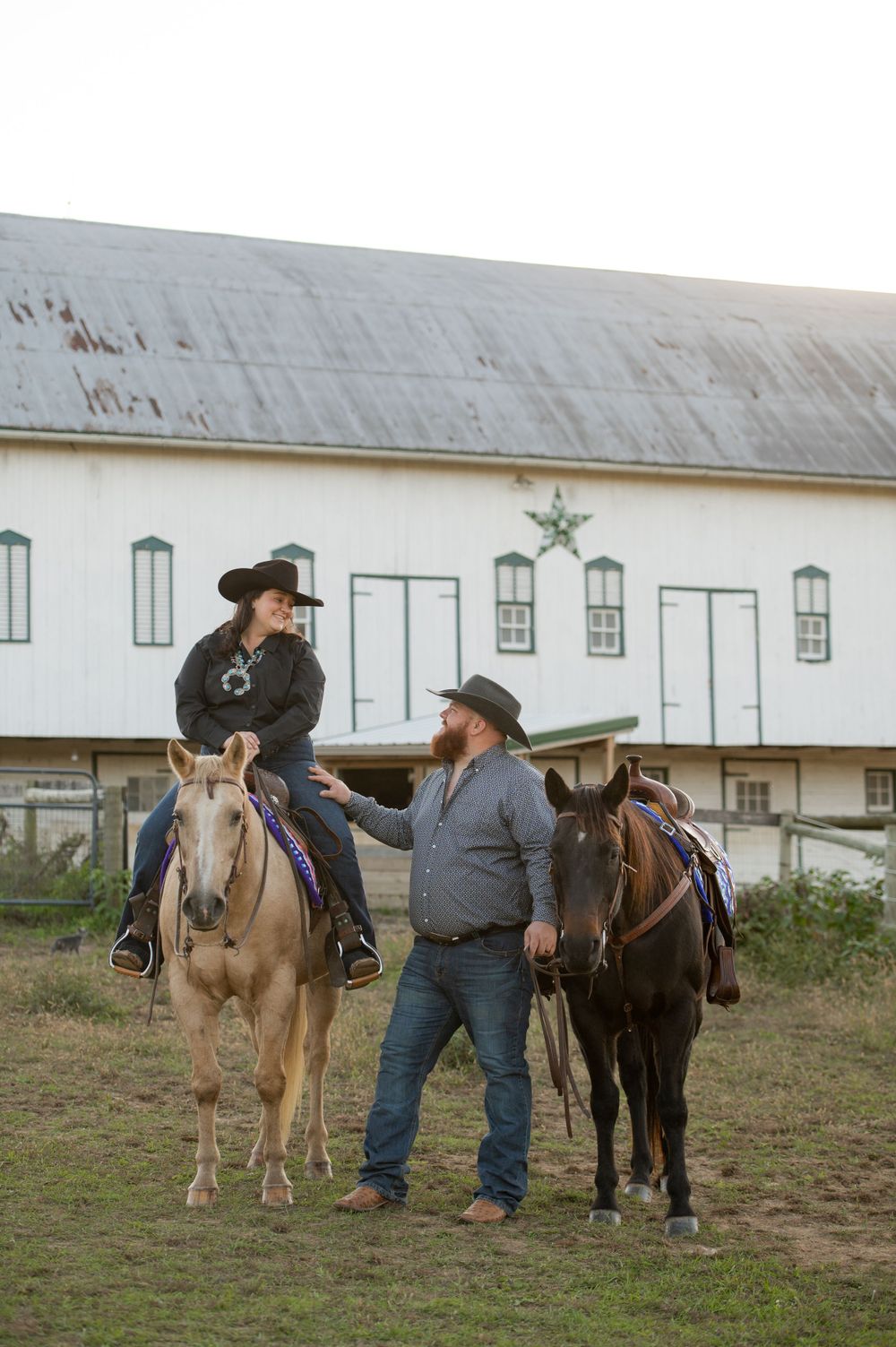 Two people with horses outside a white barn with green trim and multiple windows.