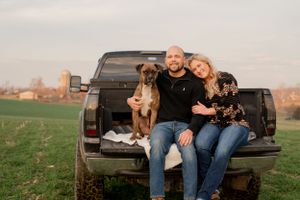 A couple sits in a pickup truck bed with their dog during a countryside photoshoot.