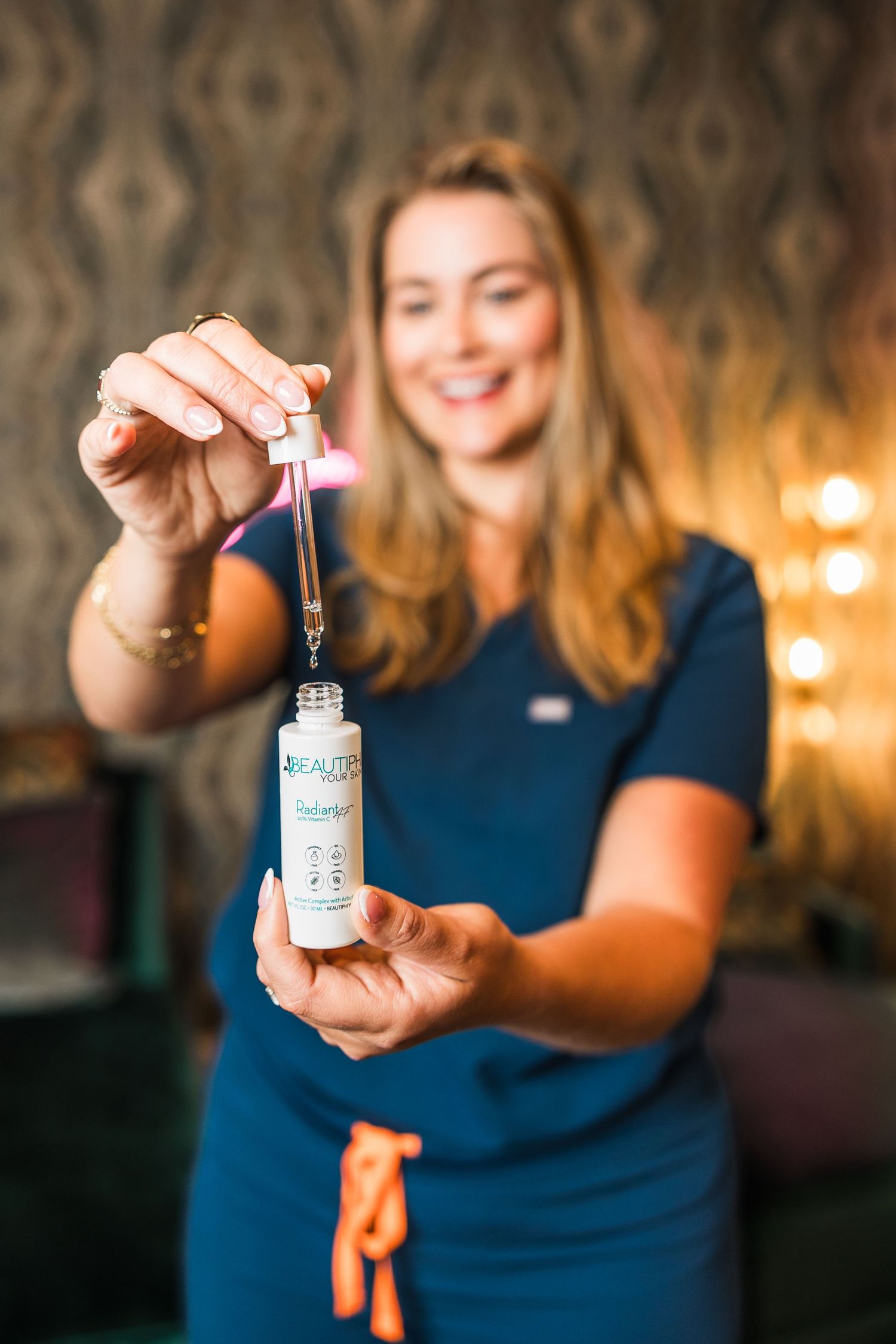 Smiling woman in blue scrubs uniform holds up a dropper bottle of Beaute skincare serum toward camera.