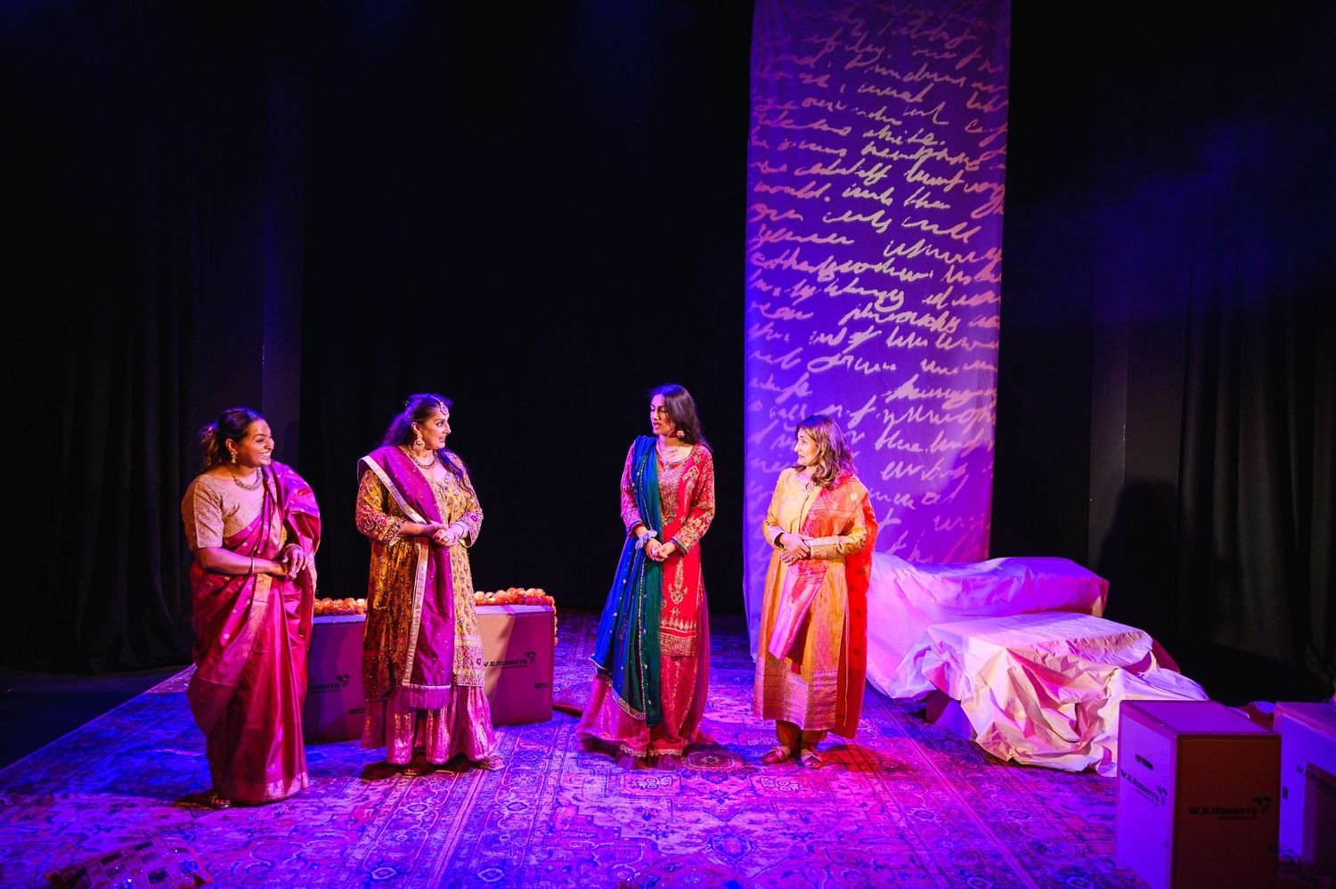 Performers in pink traditional costumes stand on stage under dramatic purple lighting.