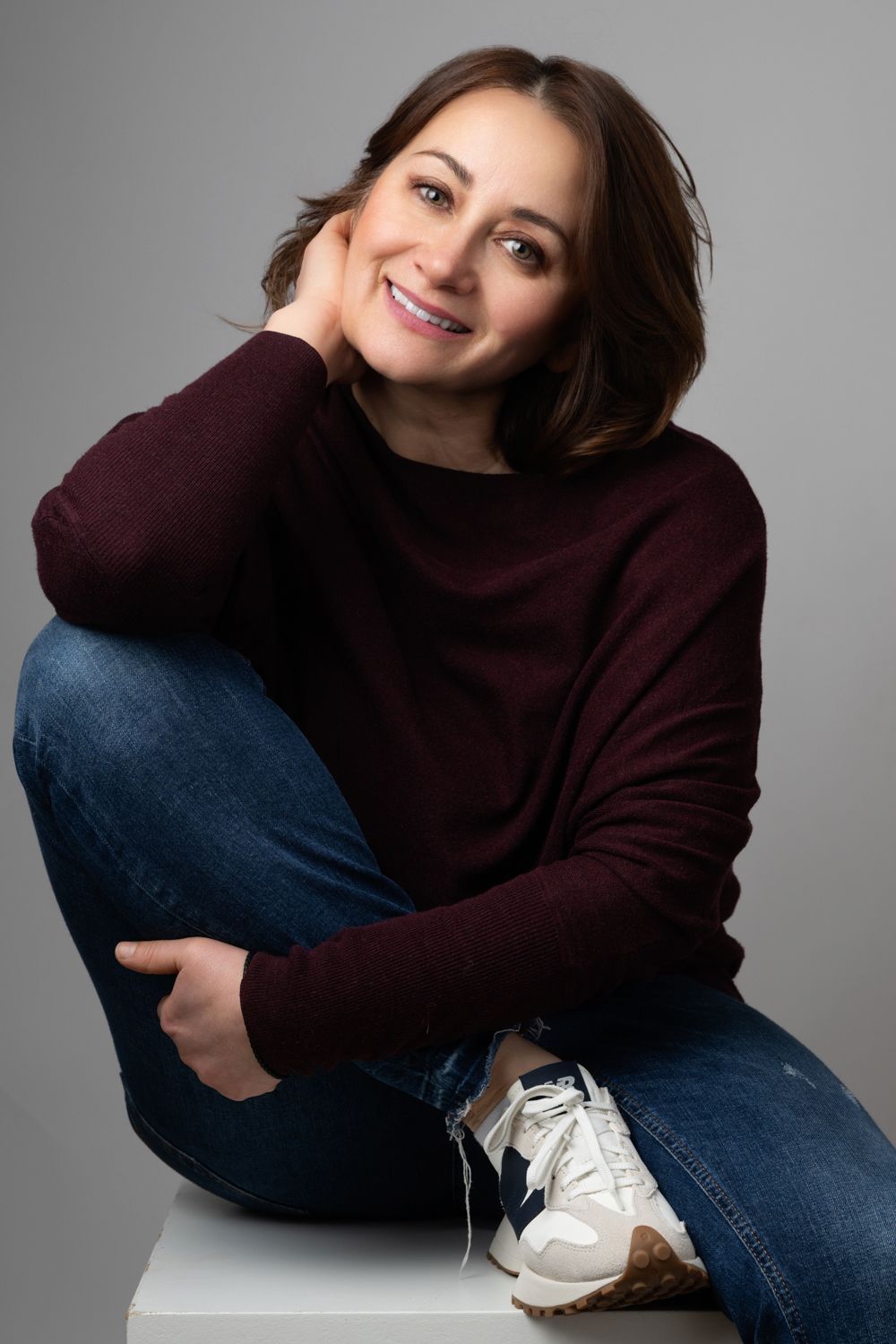 Person in burgundy sweater and jeans sits casually on white surface while smiling warmly against gray background.