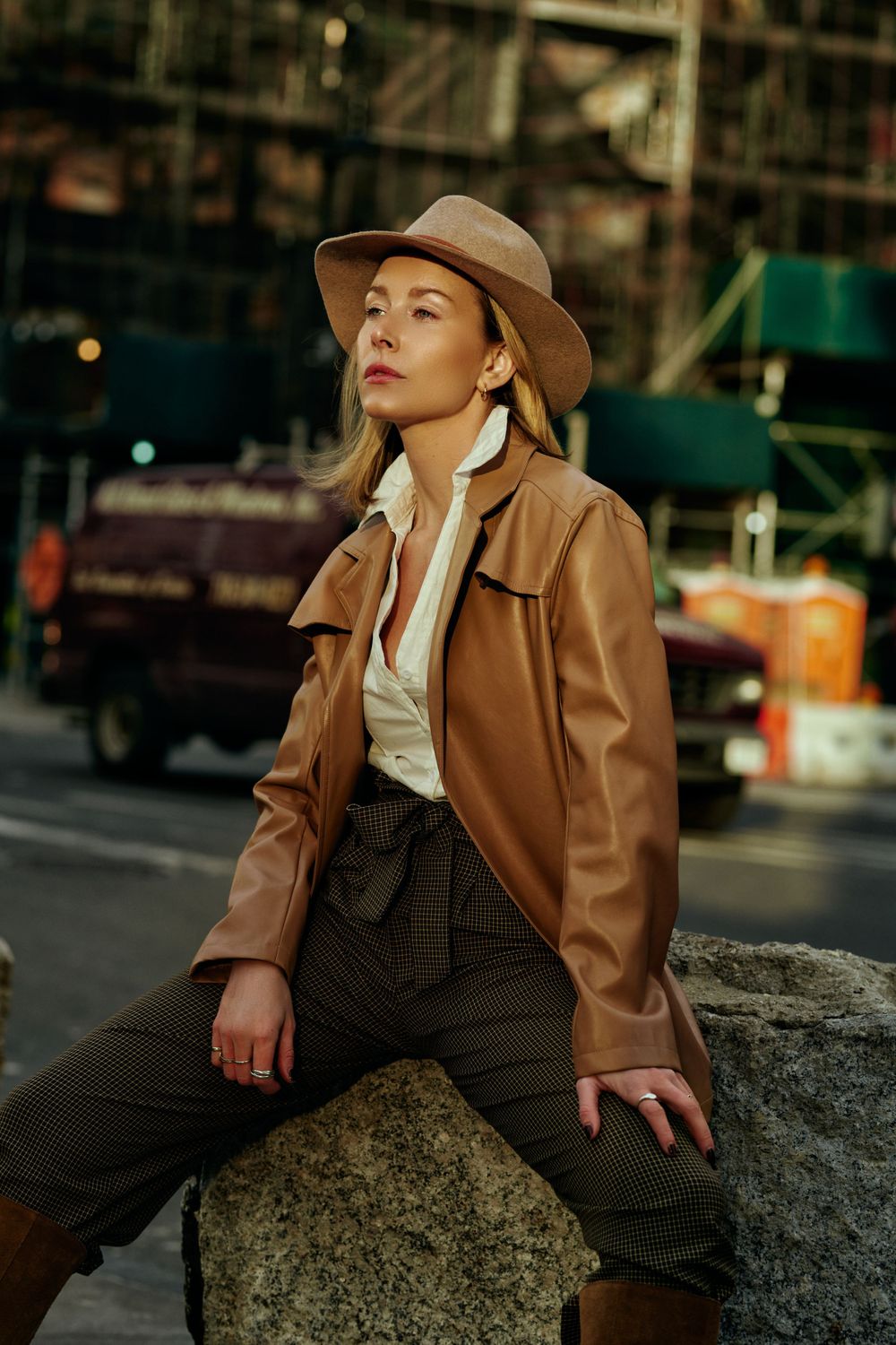 Person in brown leather jacket and hat poses on concrete structure in urban setting.