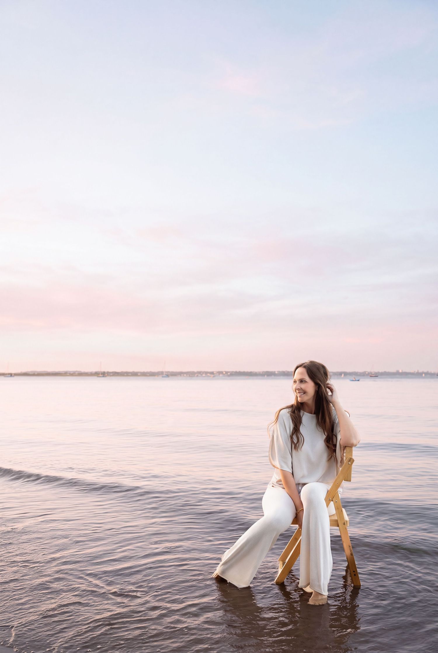 Rhiannon in a casual white attire sits on a couch holding a professional camera while smiling warmly.
