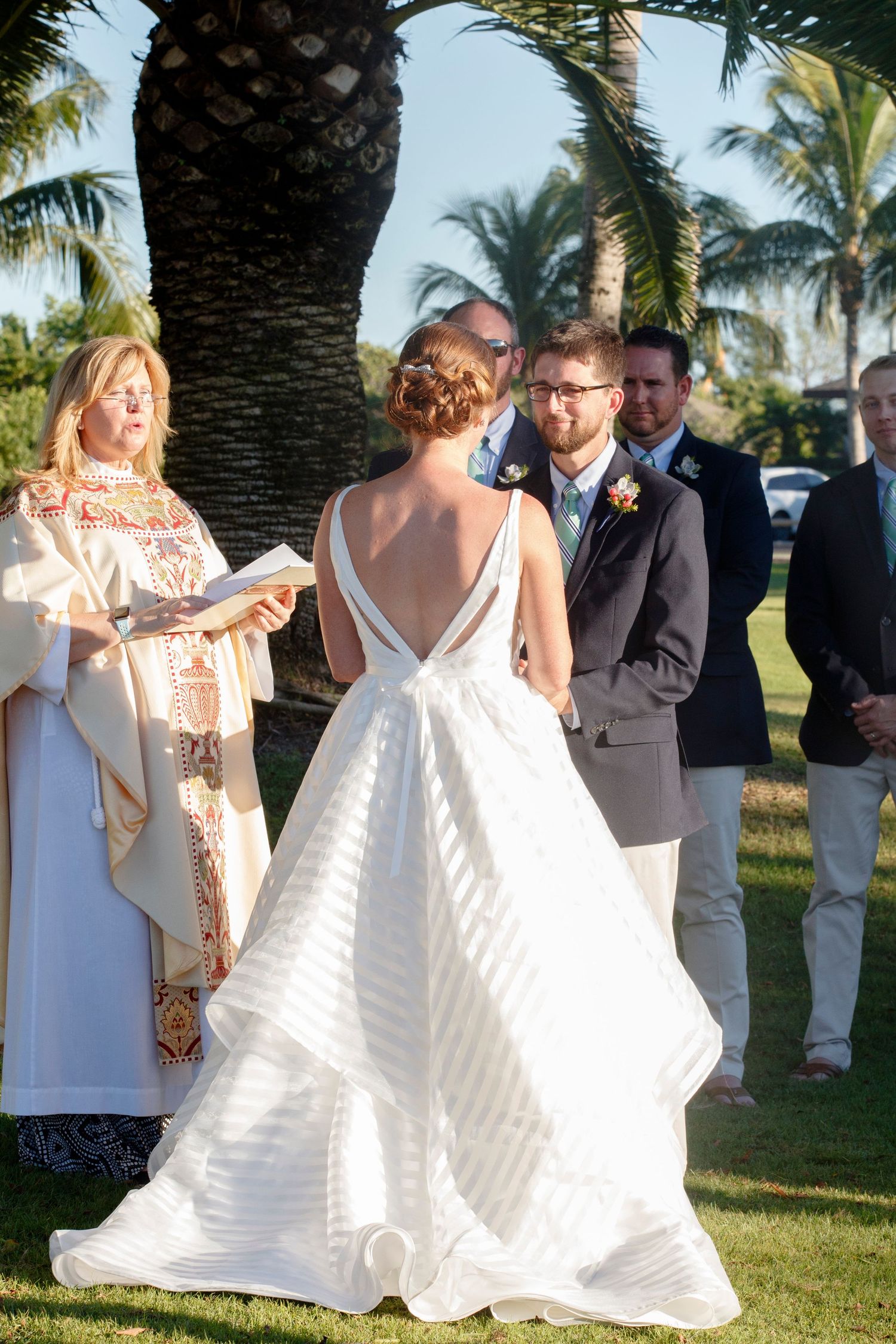 Bride in backless white striped gown exchanging vows with groom outdoors under tropical palm trees. Gasparilla Inn