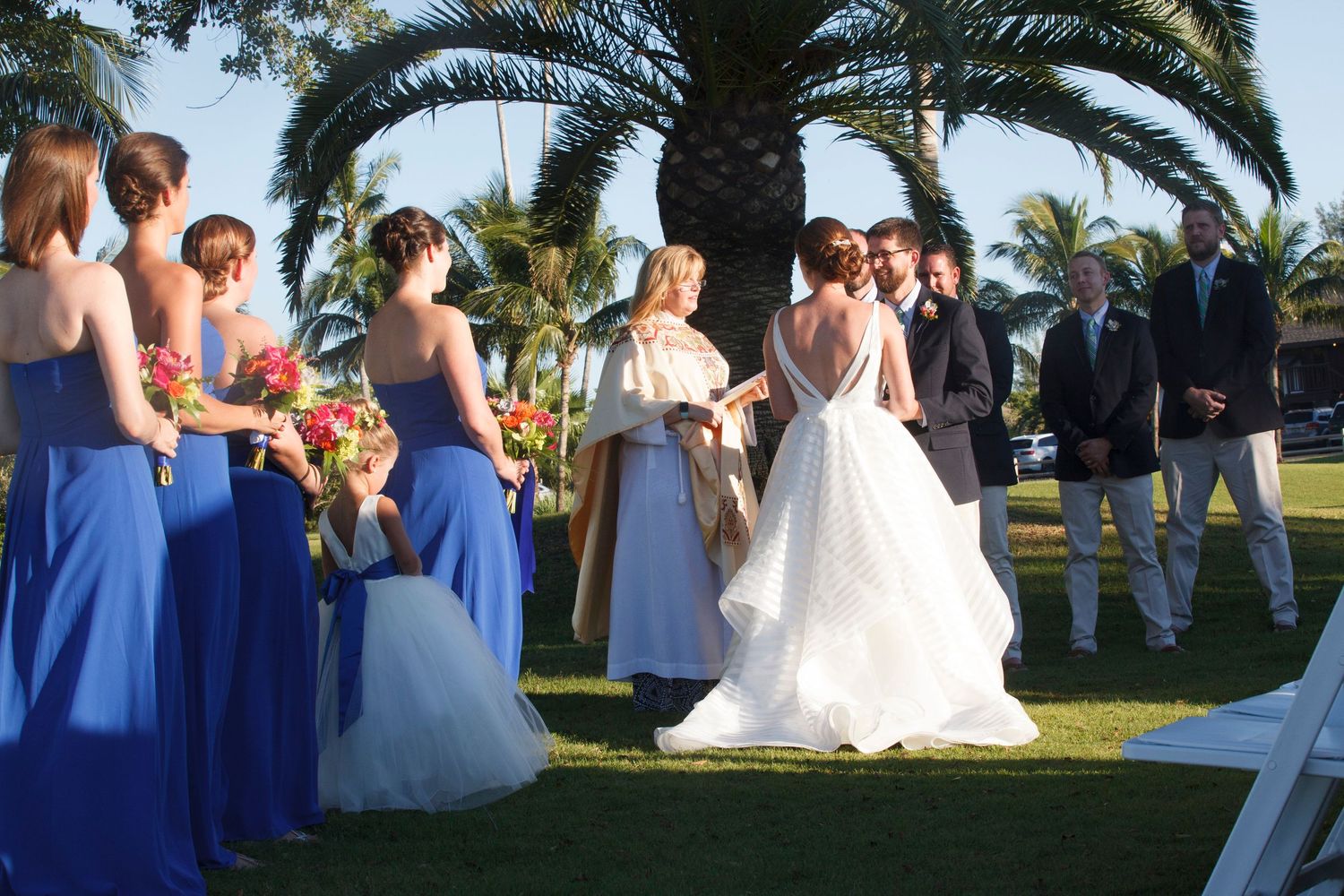Outdoor wedding ceremony with bridesmaids in blue gowns and bride in white dress under palm trees. Gasparilla Inn