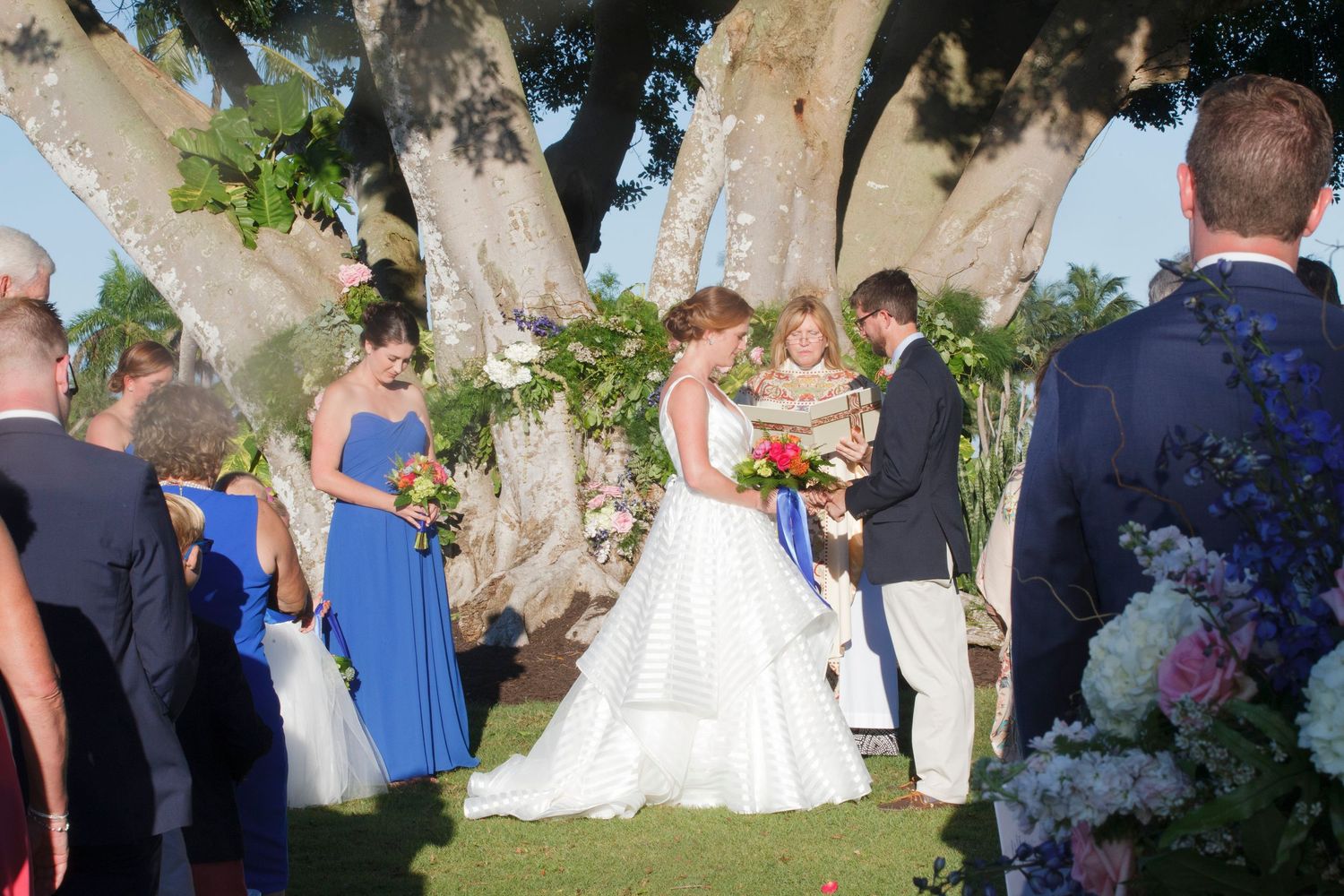 Bride and groom exchange vows beneath large tree with blue-gowned bridesmaids and floral arch. Gasparilla Inn