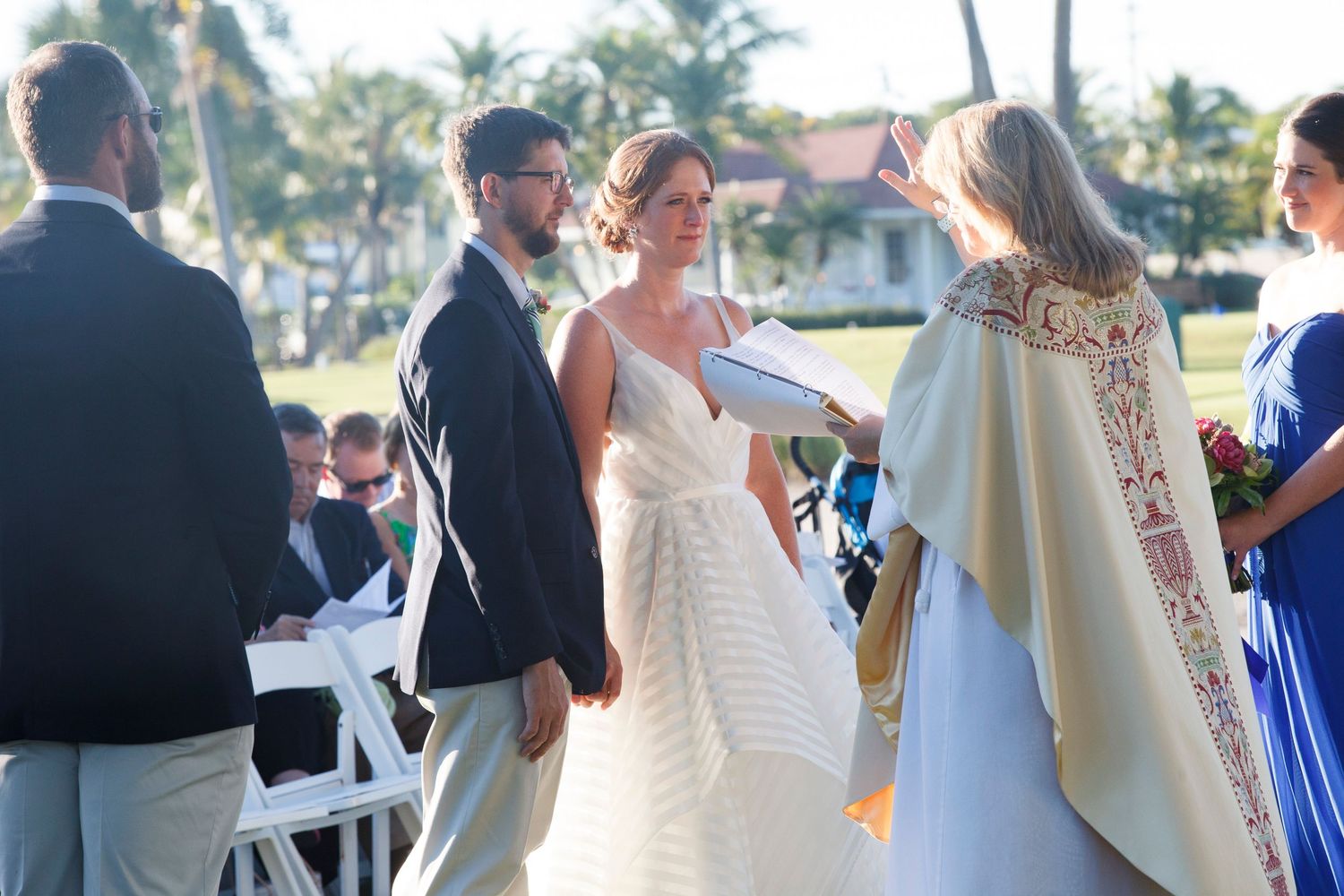 Bride in striped gown and groom hold hands as officiant in red vestments leads ceremony. Gasparilla Inn
