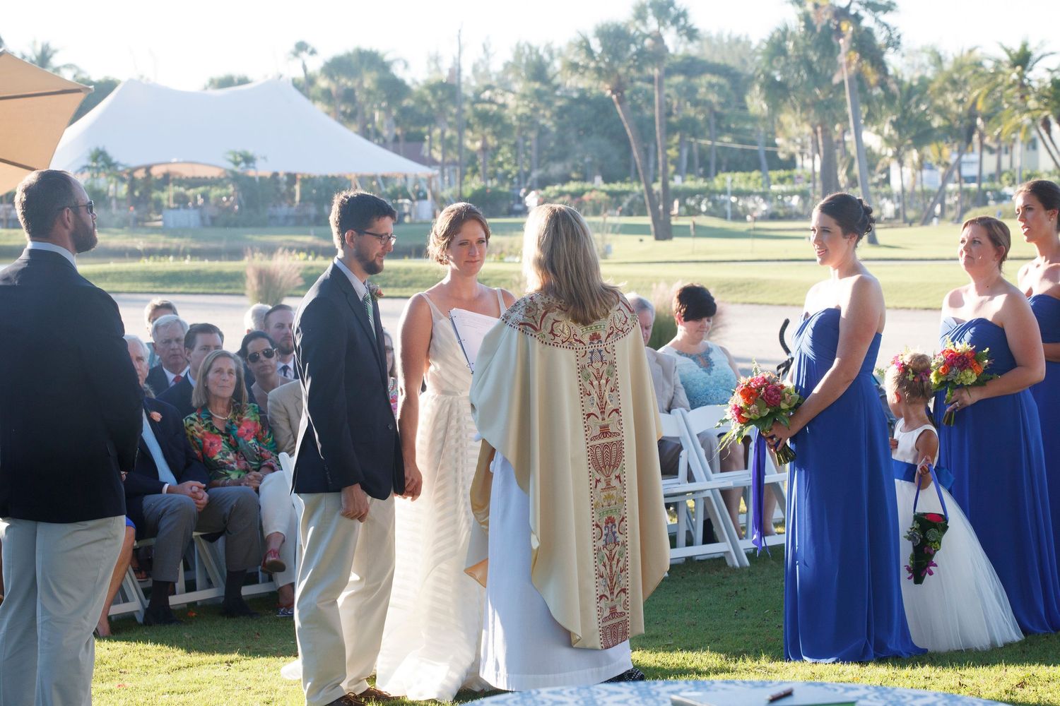 Couple exchanges vows outdoors with officiant, blue-dressed bridesmaids, and seated wedding guests. Gasparilla Inn