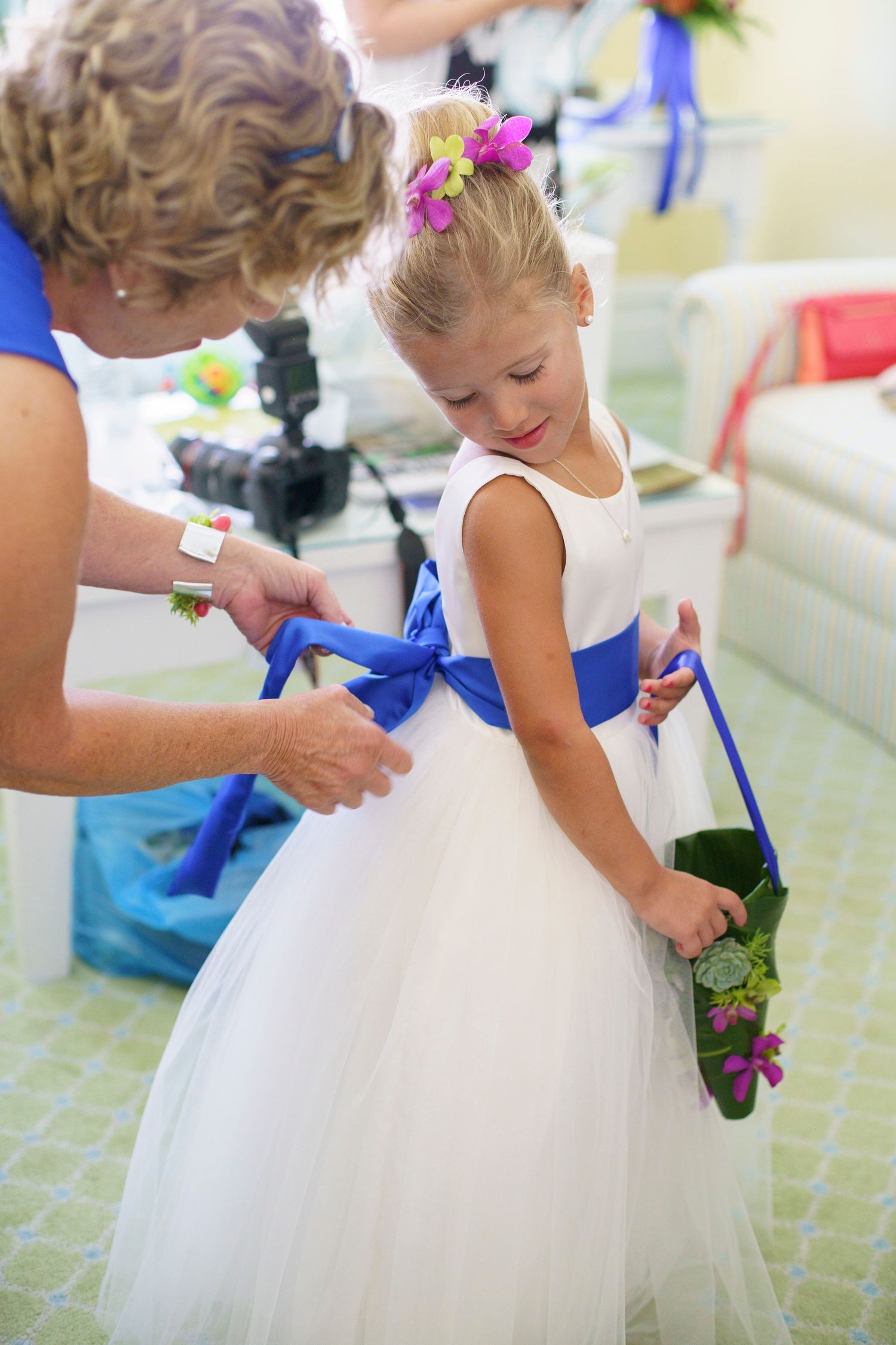Woman ties blue sash on flower girl in white tulle dress holding floral basket. Gasparilla Inn