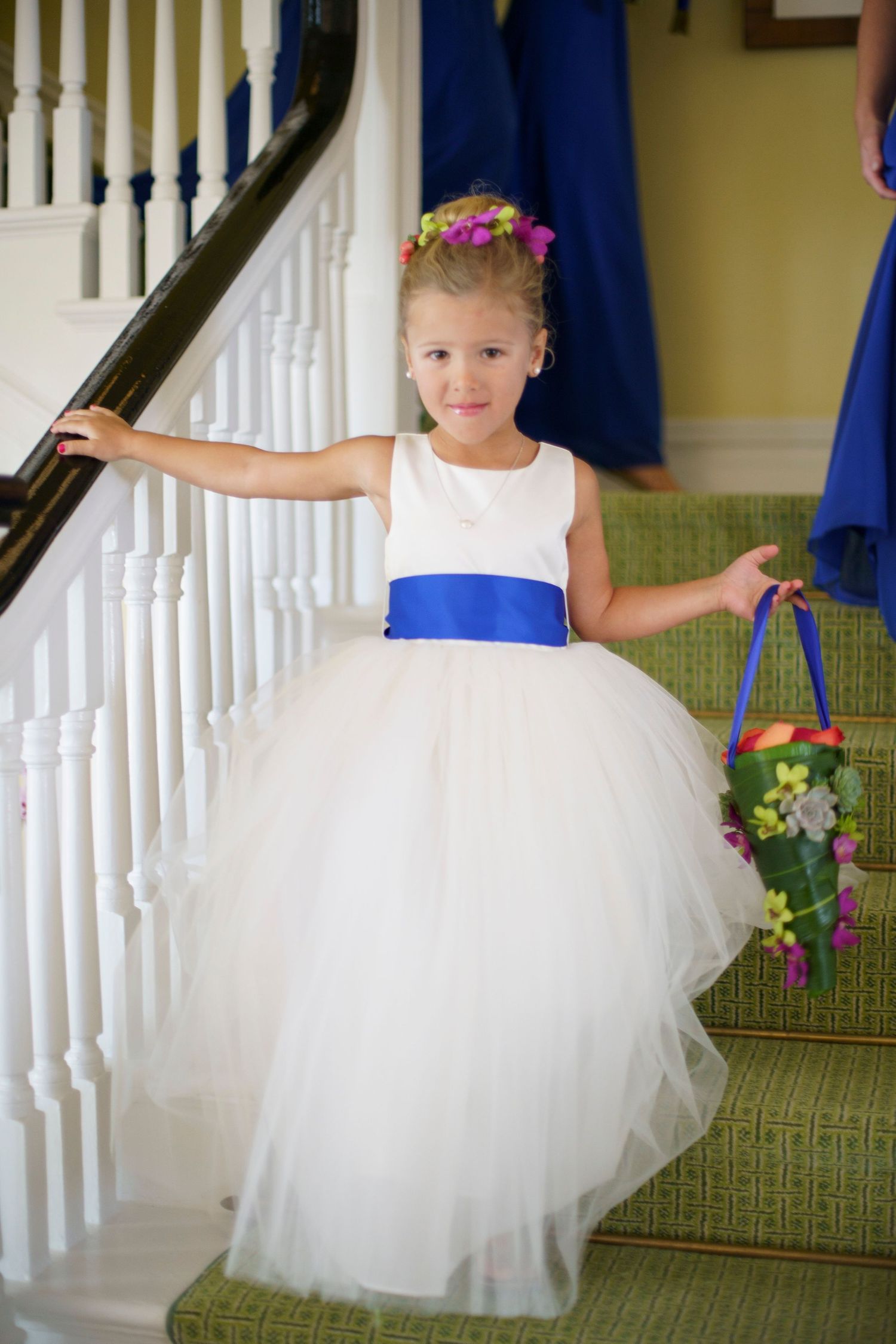 Flower girl in white tulle dress with blue sash carries floral basket down staircase. Gasparilla Inn