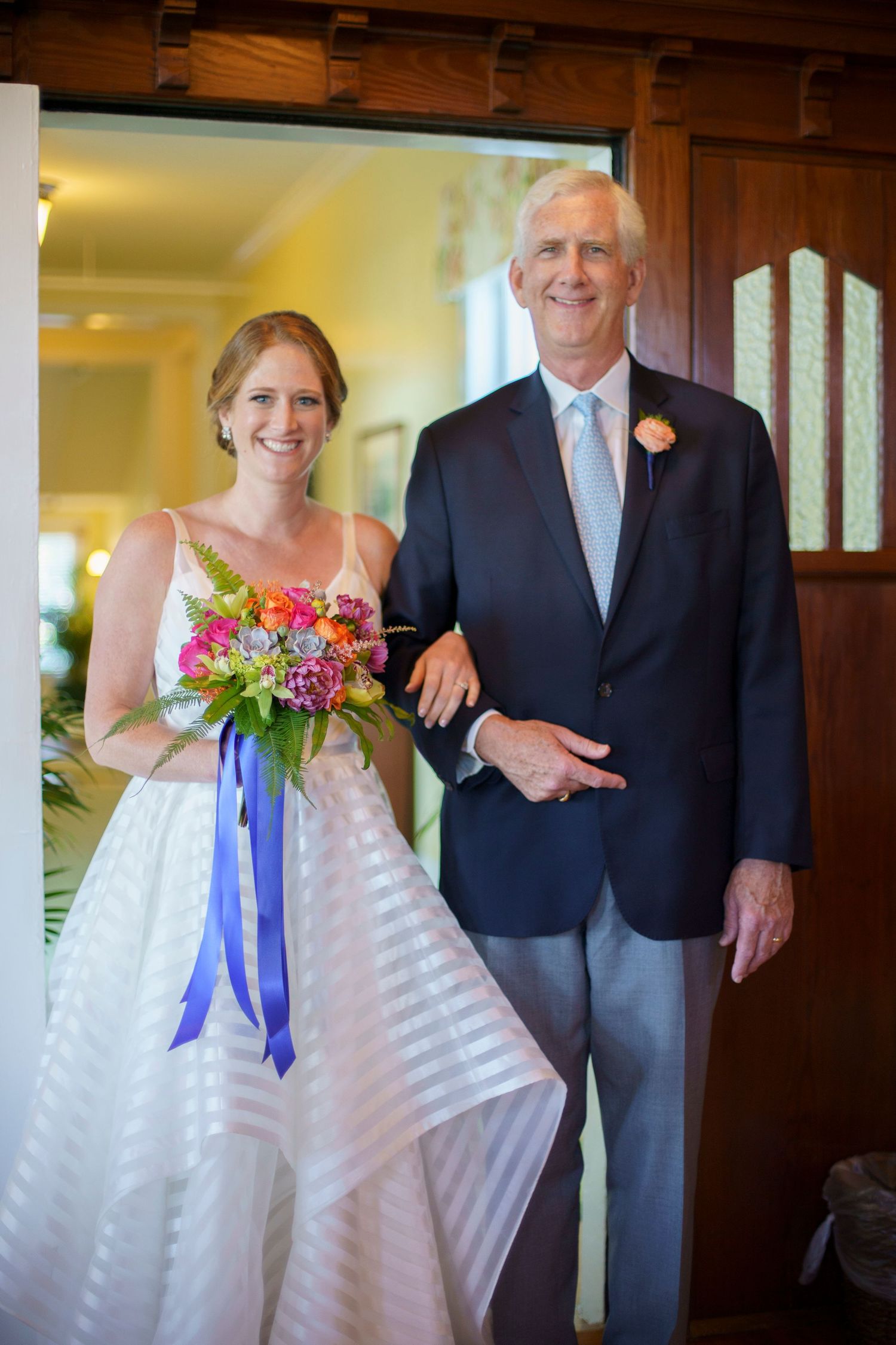 Bride in striped gown holds colorful bouquet with blue ribbon beside escort at doorway. Gasparilla Inn