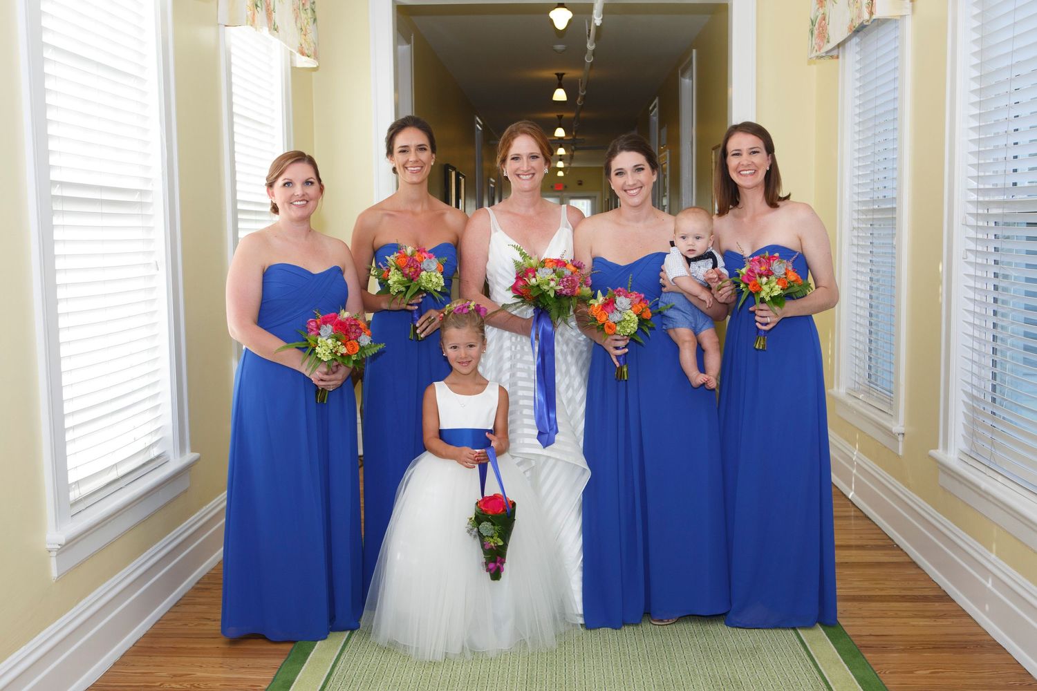 Bride with bridesmaids in royal blue gowns holding vibrant bouquets in hallway. Gasparilla Inn