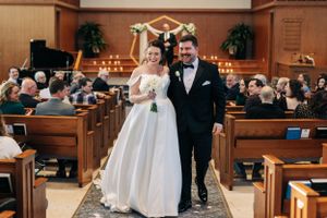 A couple walks down the church aisle during their wedding ceremony as guests watch from wooden pews.