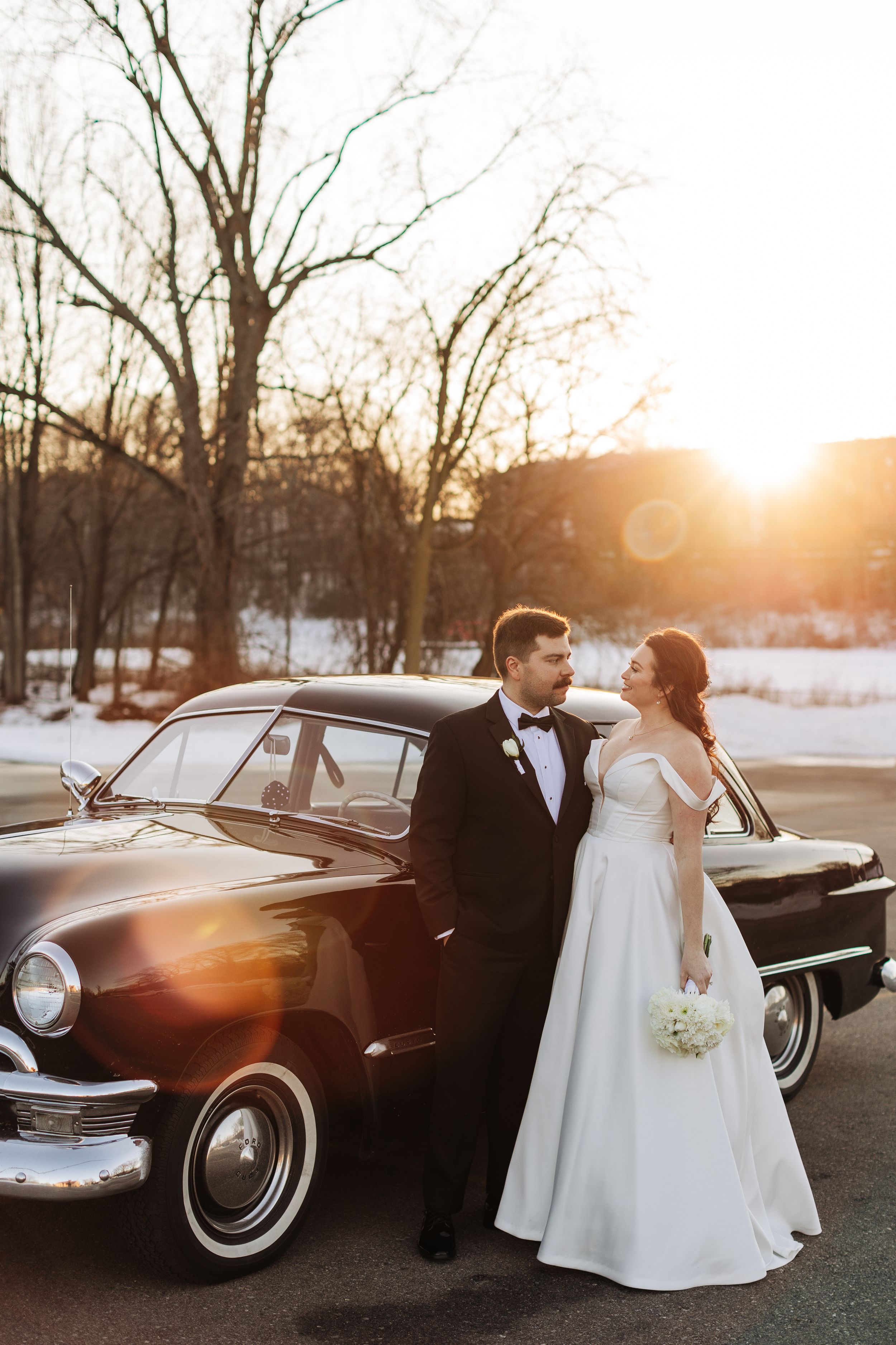 A bride and groom pose with a vintage black car at sunset in a snowy winter landscape.