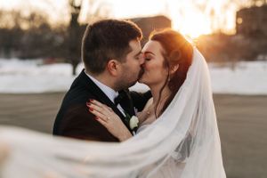 Romantic wedding portrait of a couple kissing during golden hour sunset with flowing white veil in foreground.