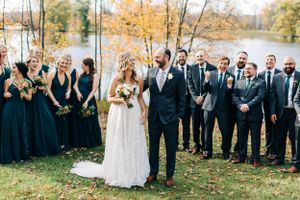 A wedding party in navy and black attire poses together by a lake in autumn.