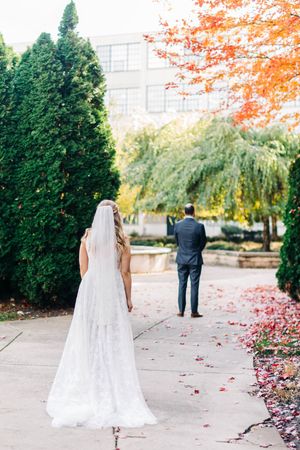 A bride in a flowing white gown waits as her partner stands ahead on a fall-colored path.