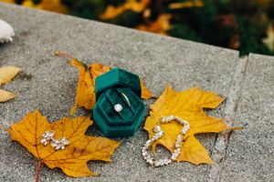 Wedding accessories including a green flower and jewelry arranged on fall leaves against concrete.