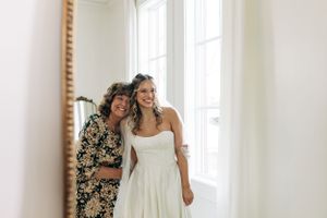 Two people share a joyful moment together in a sunlit room beside a window, one wearing a floral dress and one in white.