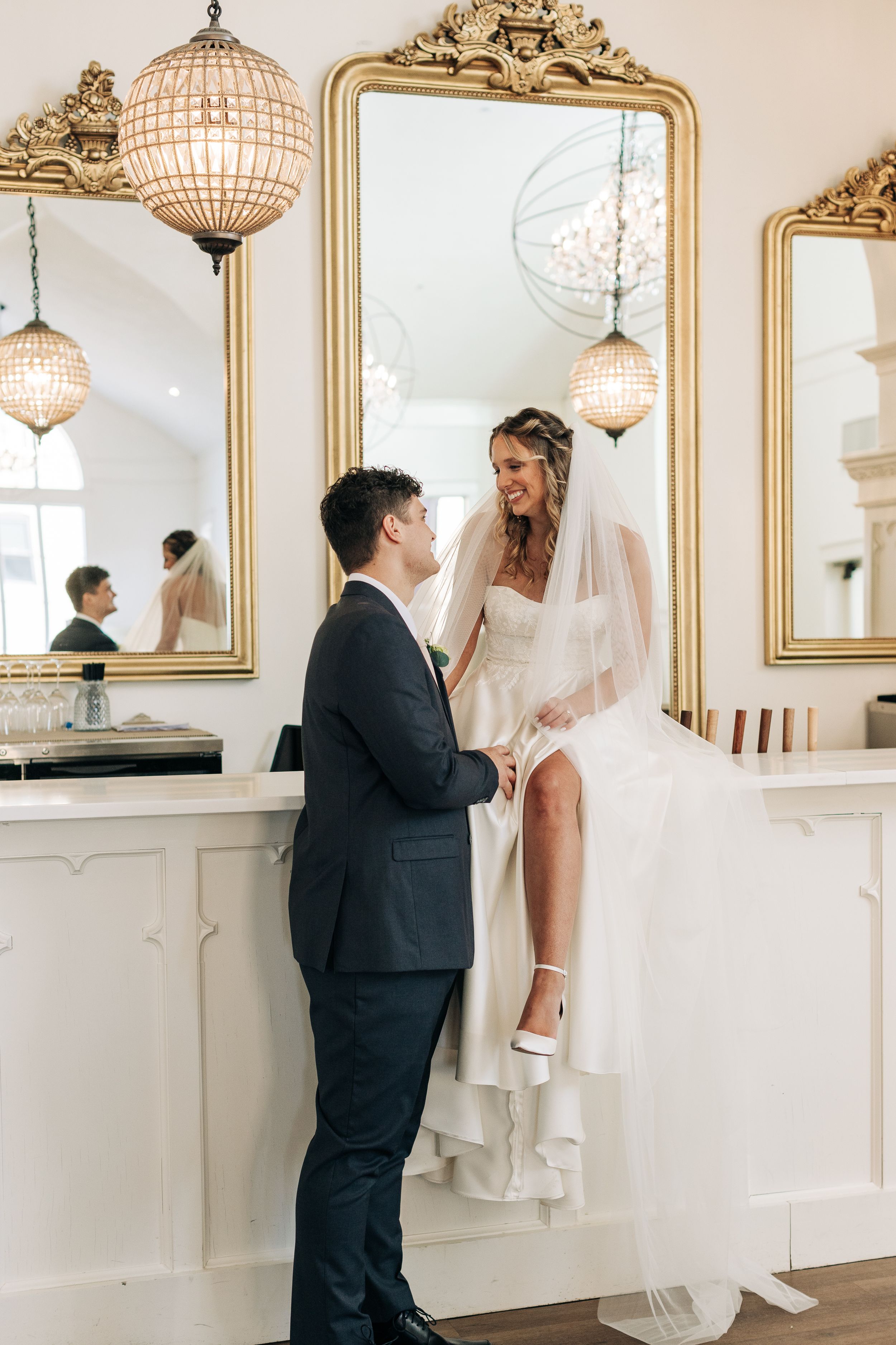 Couple shares intimate moment in elegant venue with ornate gold mirrors and crystal chandeliers during wedding photoshoot.