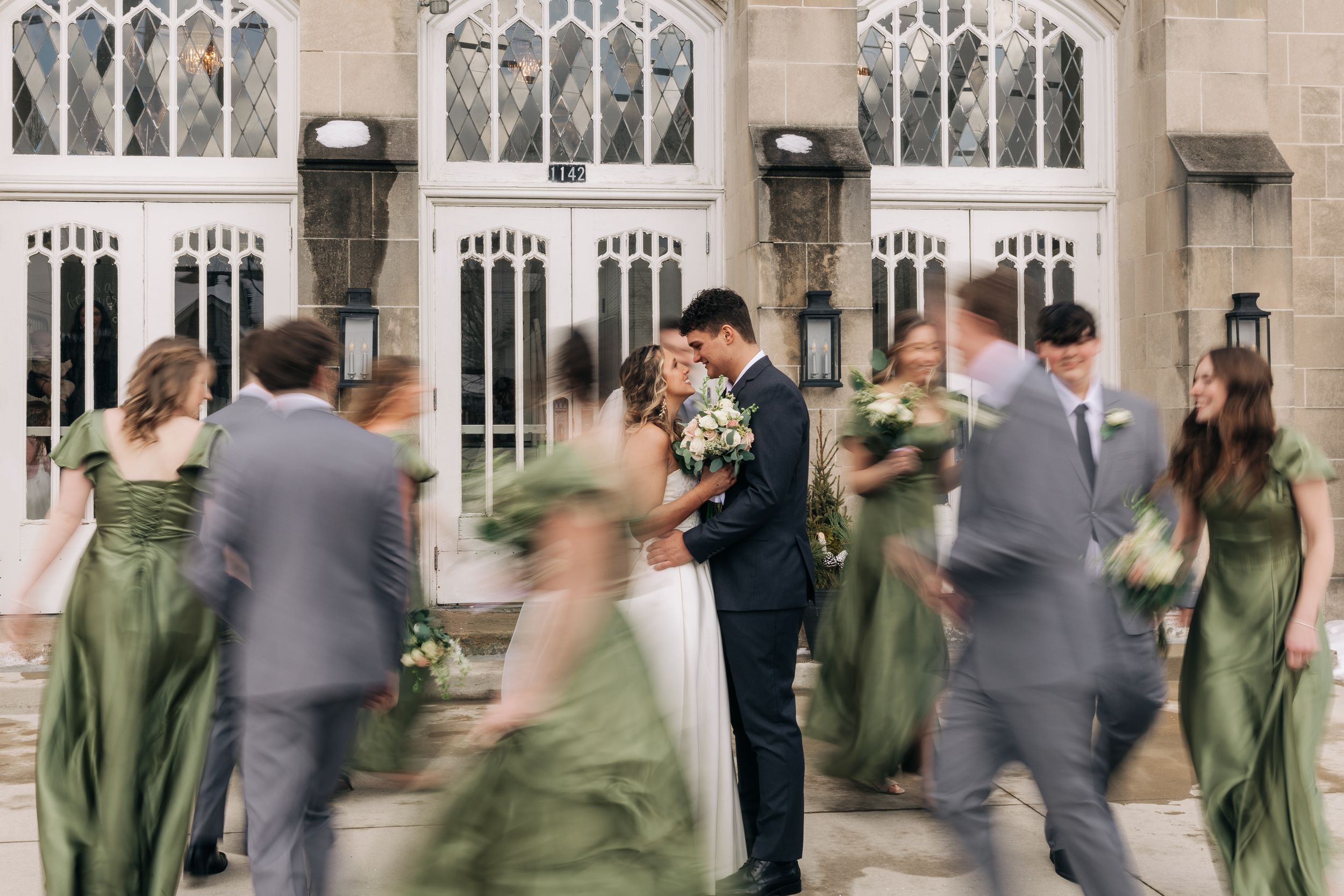 Wedding party wearing sage green dresses and gray suits stands in front of a gothic style building during a ceremony.