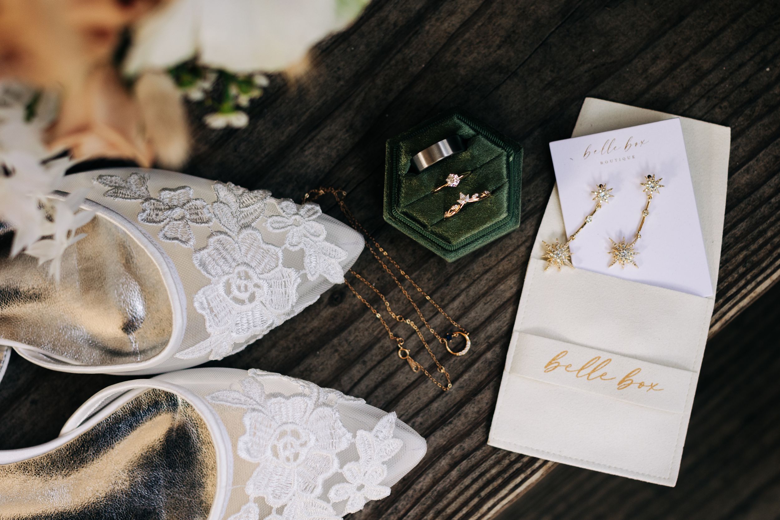 Wedding details including lace shoes, jewelry, and stationery arranged on wooden surface.