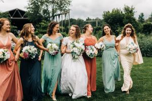Wedding party in multicolored bridesmaid dresses walking across a garden lawn with bouquets.