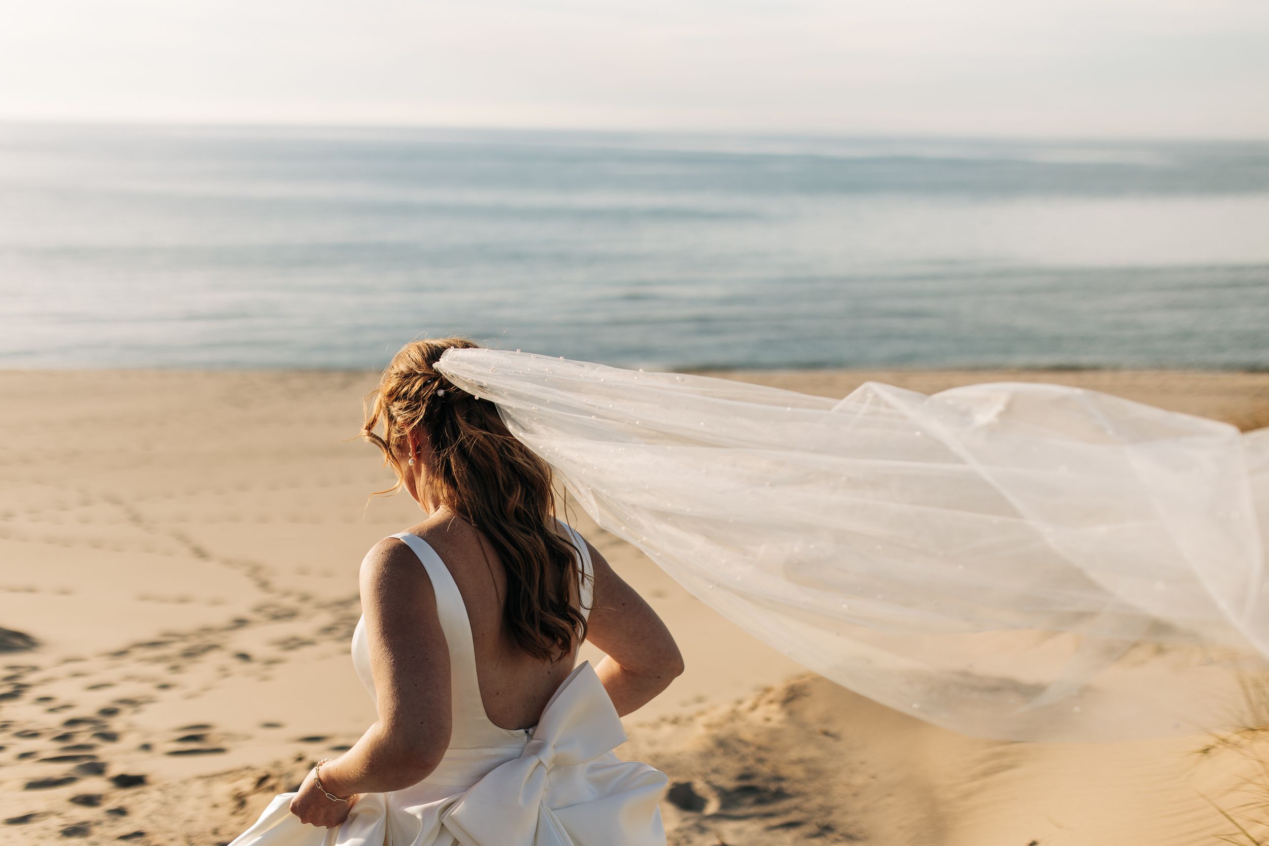 A bride's veil flows in the wind on a sandy beach with ocean views.