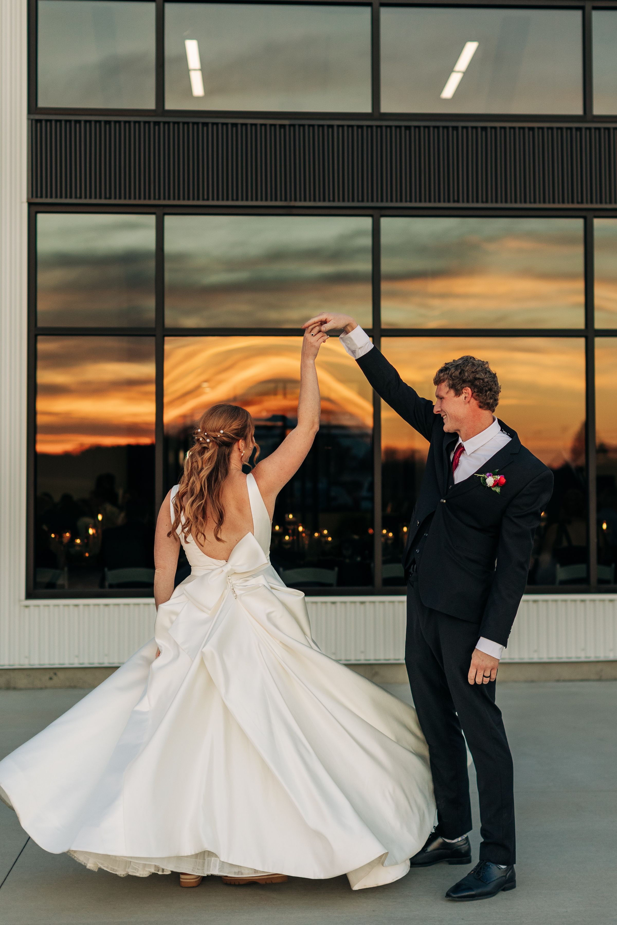 A newlywed couple dances outside a modern building during a vibrant sunset.