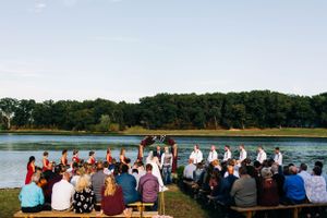 An outdoor lakeside wedding ceremony with guests seated on wooden benches facing the water.