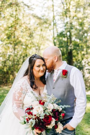 A romantic wedding portrait taken in a sunlit wooded setting with a red and white bouquet.