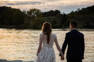 Newlyweds hold hands while walking along waterfront at golden hour.