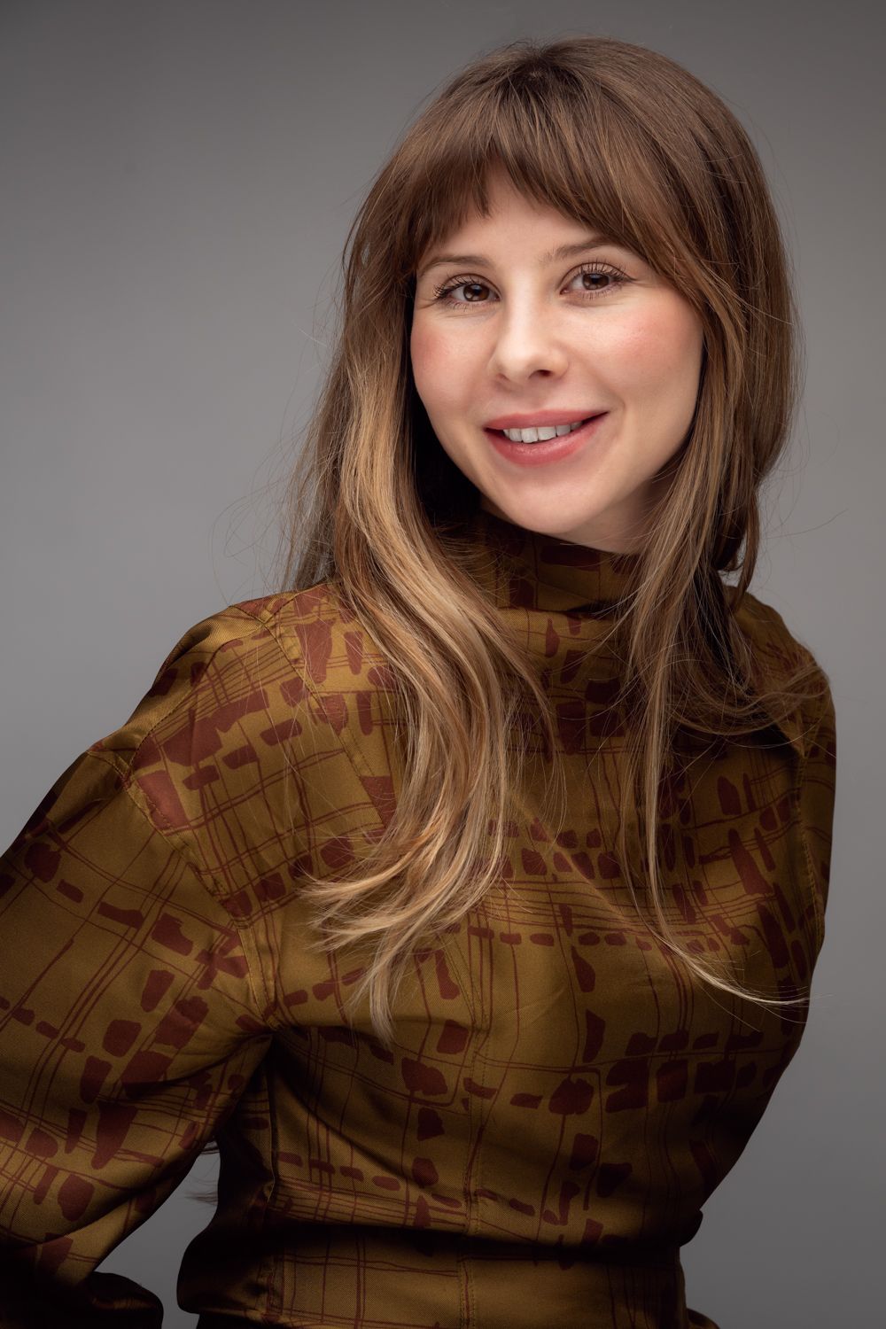 Professional portrait of someone wearing a brown patterned turtleneck sweater against a gray background.