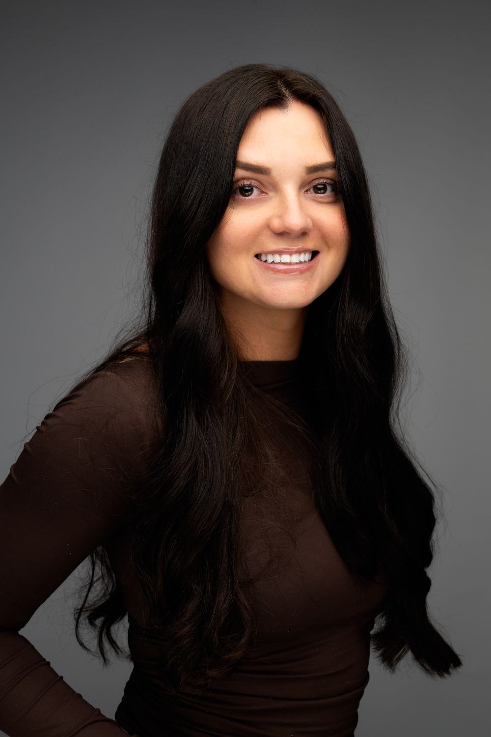 Studio portrait of a person with long dark hair wearing a brown long-sleeve top against a gray background.