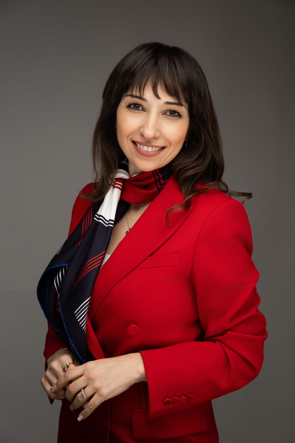 Professional portrait of someone wearing a bright red blazer and striped scarf against a grey background.